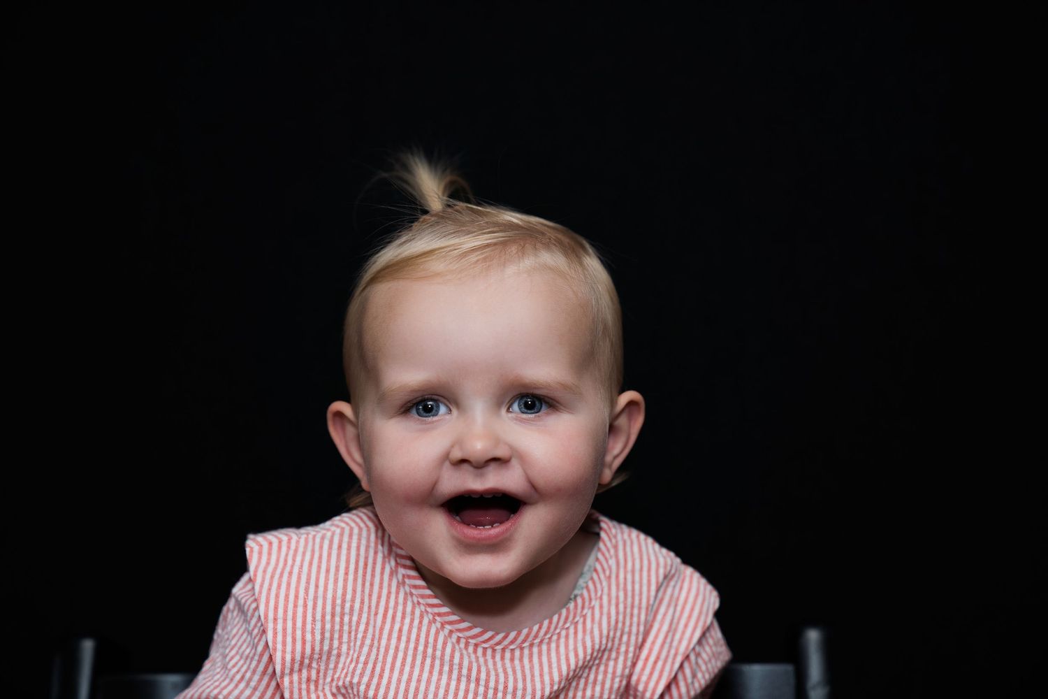 Een vrolijk lachend babymeisje met een klein staartje, gekleed in een roze gestreept pakje, op een zwarte achtergrond.
