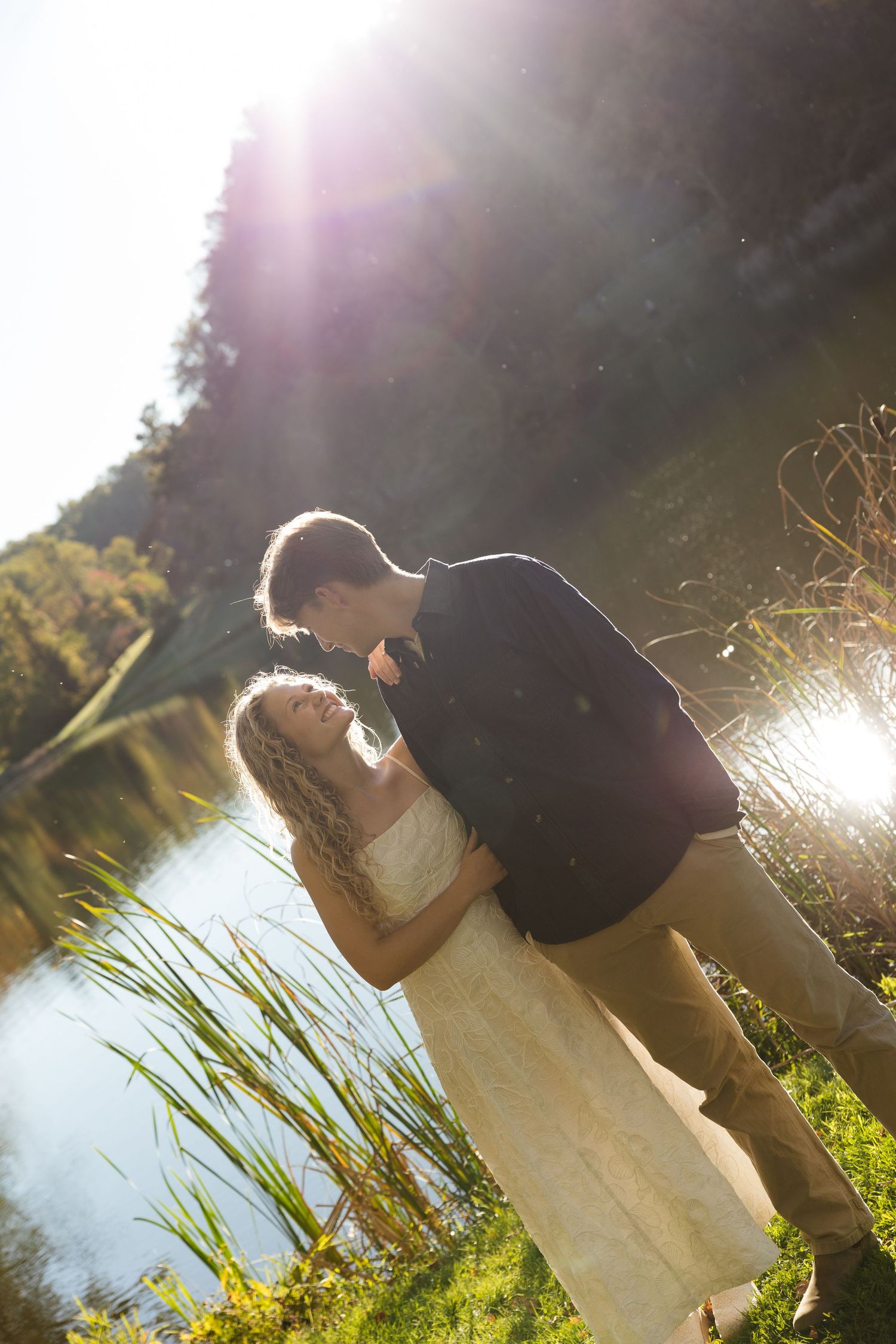 Black and white photo of a couple holding hands by a sunlit lake, woman in white dress, romantic outdoor setting.