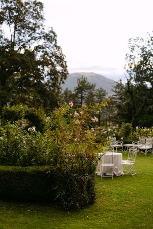 Romantic outdoor wedding reception setup with white tables on green lawn surrounded by mountains and lush gardens.