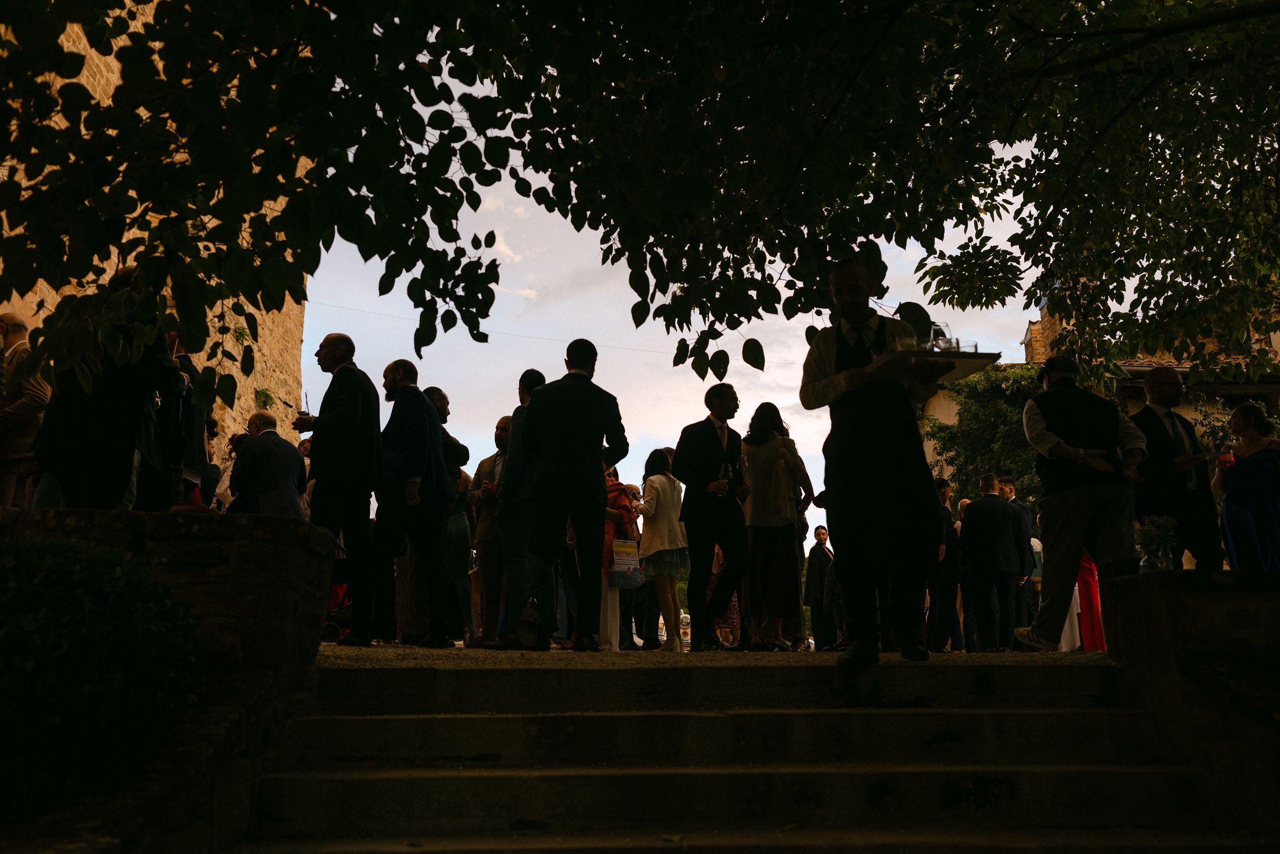 Silhouettes of wedding guests gathered under trees at dusk during an outdoor wedding celebration.