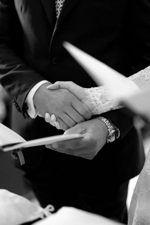 Black and white close-up of bride and groom holding hands during a wedding ceremony signing.