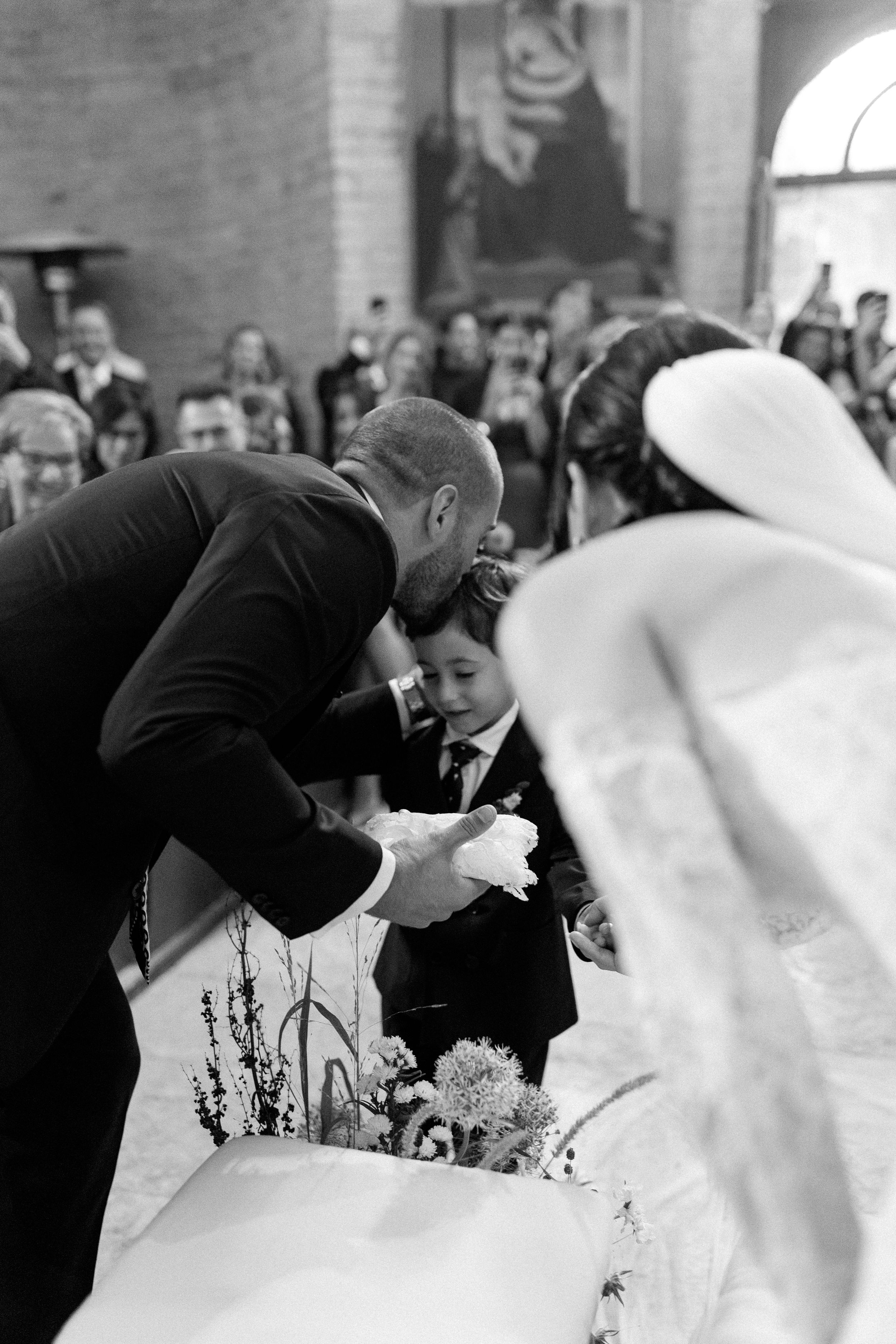 Black and white photo of groom kneeling to embrace young boy during wedding ceremony at altar.
