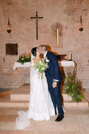 Bride and groom share first kiss at rustic stone church altar with floral arrangements.