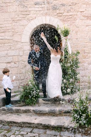 Joyful bride and groom exit stone church doorway as guests throw rice confetti.