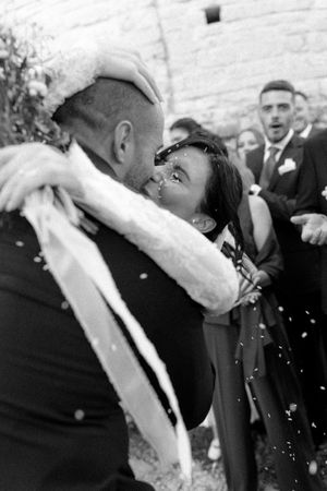 Joyful newlyweds share a kiss surrounded by confetti and wedding guests in black and white photo.