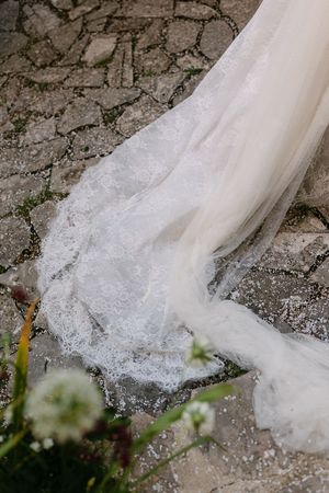 Close-up of elegant lace wedding dress train trailing on cobblestone ground with white flowers.