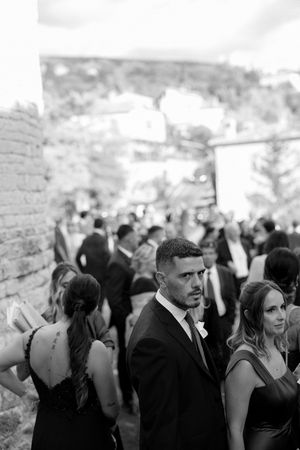 Elegant wedding guest in suit looking back at camera amid crowd at outdoor reception.