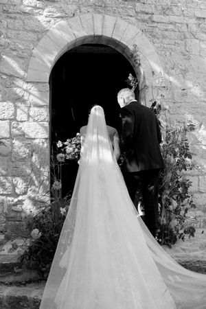 Black and white photo of bride in lace gown and veil entering stone church with escort.