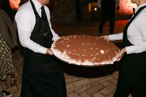Wedding servers carrying large tiramisu dessert on tray at outdoor evening reception.