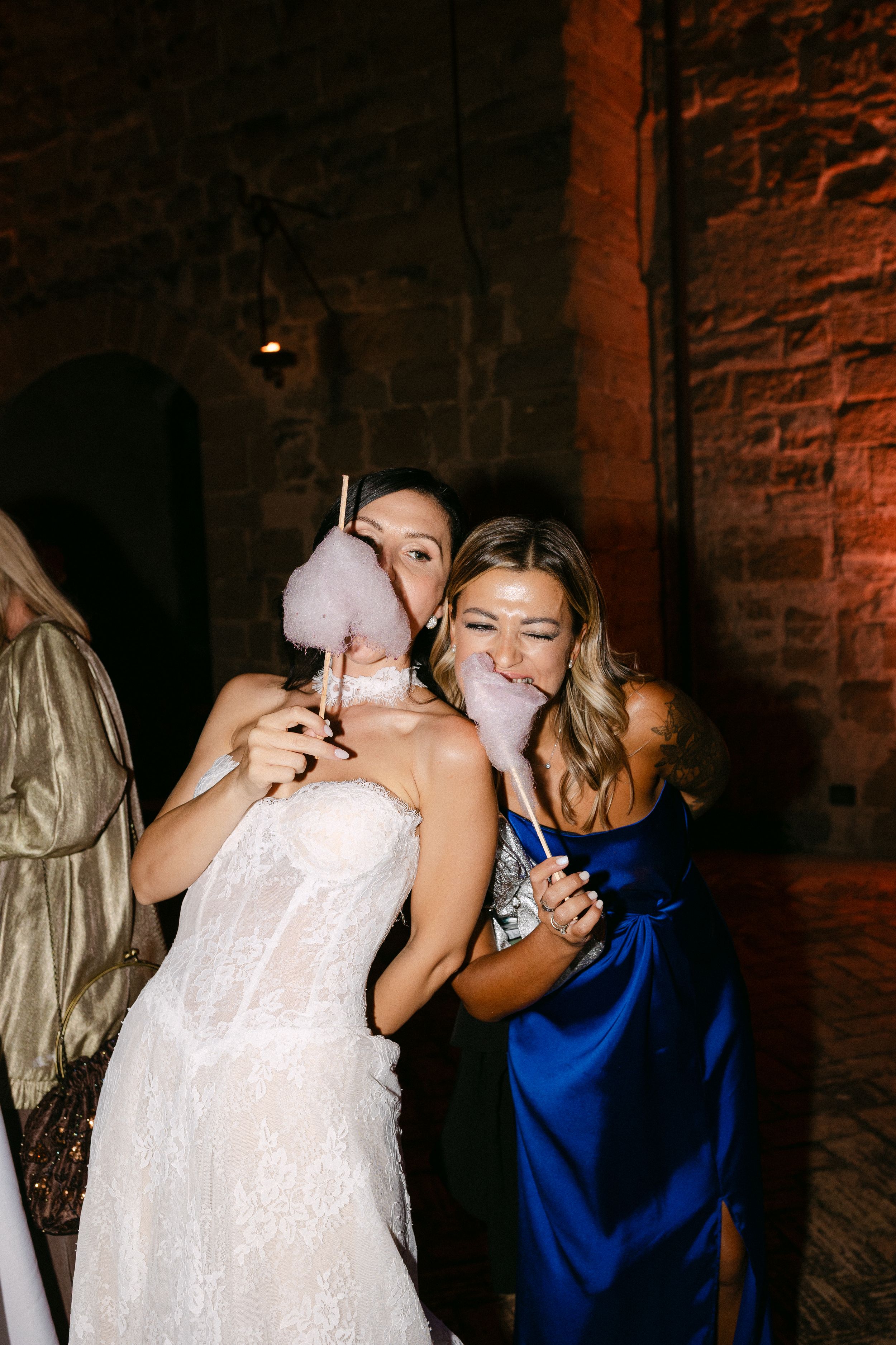 Bride in lace gown and bridesmaid in blue satin dress share cotton candy at a rustic stone venue wedding reception.