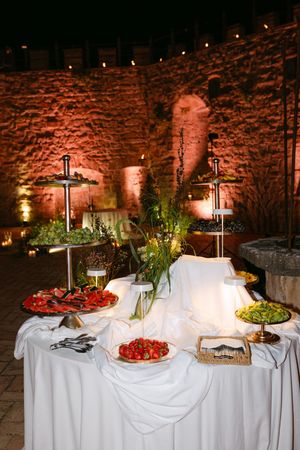 Elegant outdoor wedding buffet table with fresh fruits and appetizers against illuminated stone wall.