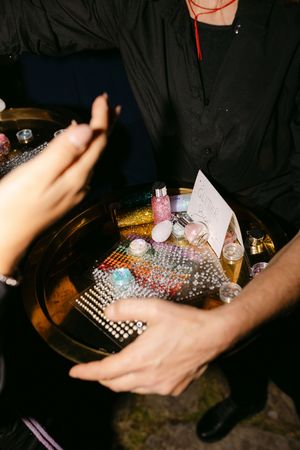 Person in black shirt holding a gold tray with glitter, rhinestones, and beauty accessories at an event.