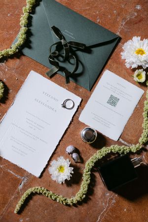Flat lay of wedding stationery suite with dark envelope, invitation cards, rings, and white flowers on terracotta.
