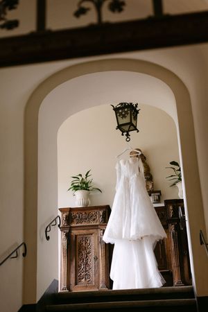 White wedding dress hanging in arched doorway of elegant venue with ornate wooden furniture and lantern.