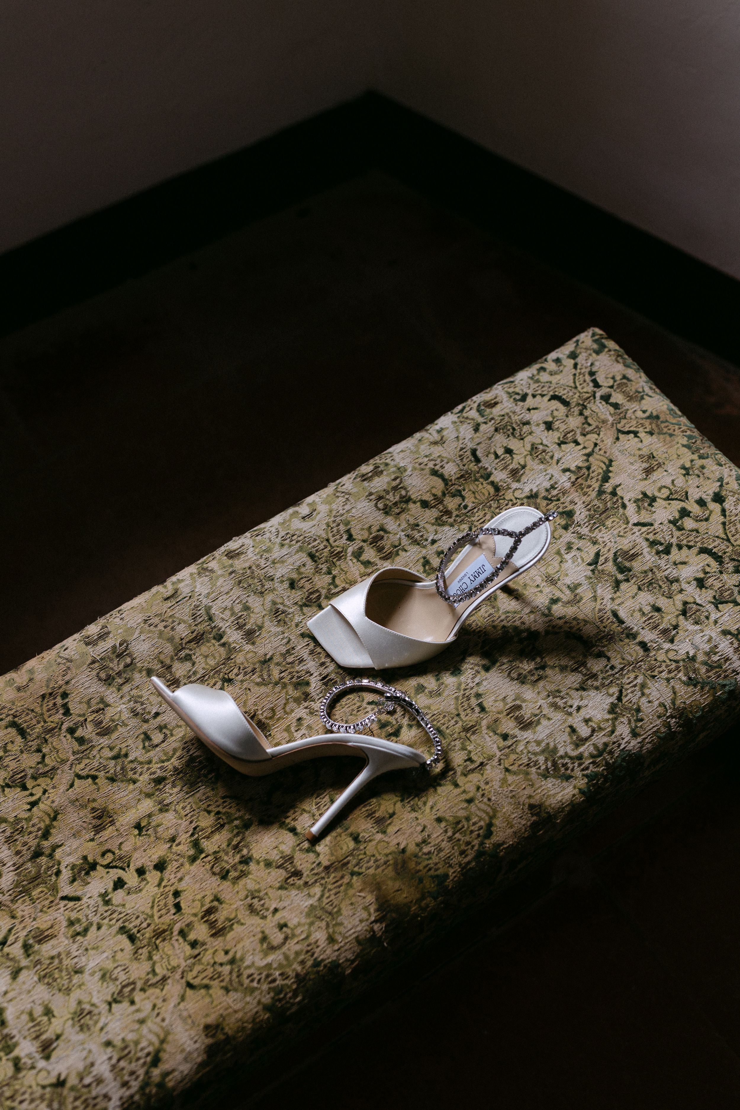 Elegant Jimmy Choo bridal heels with crystal straps resting on ornate upholstered bench in dim light.
