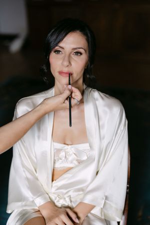 Bride in silk robe gets lip makeup applied by artist during wedding preparation indoors.