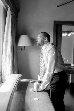 Groom in white shirt and tie gazes thoughtfully out window in black and white getting ready photo.
