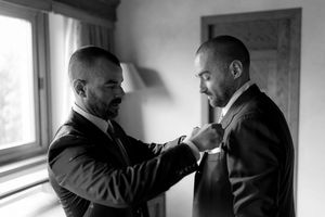 Black and white photo of a man adjusting the boutonniere on a groom's suit lapel before the wedding.