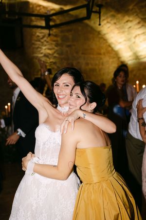 Bride in white lace gown joyfully hugging bridesmaid in gold dress at reception.