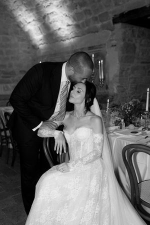 Black and white photo of groom tenderly kissing bride on head at candlelit reception table in stone venue.