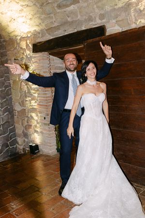 Joyful newlyweds making grand entrance into rustic stone-walled Italian wedding reception venue.