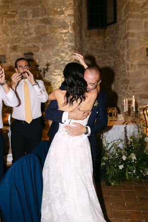 Groom in navy suit shares first dance embrace with bride in lace gown in stone venue.