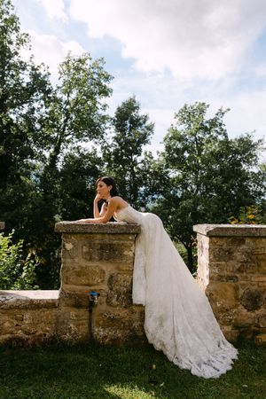 Bride in lace wedding gown leans on stone wall surrounded by lush green trees and cloudy sky.