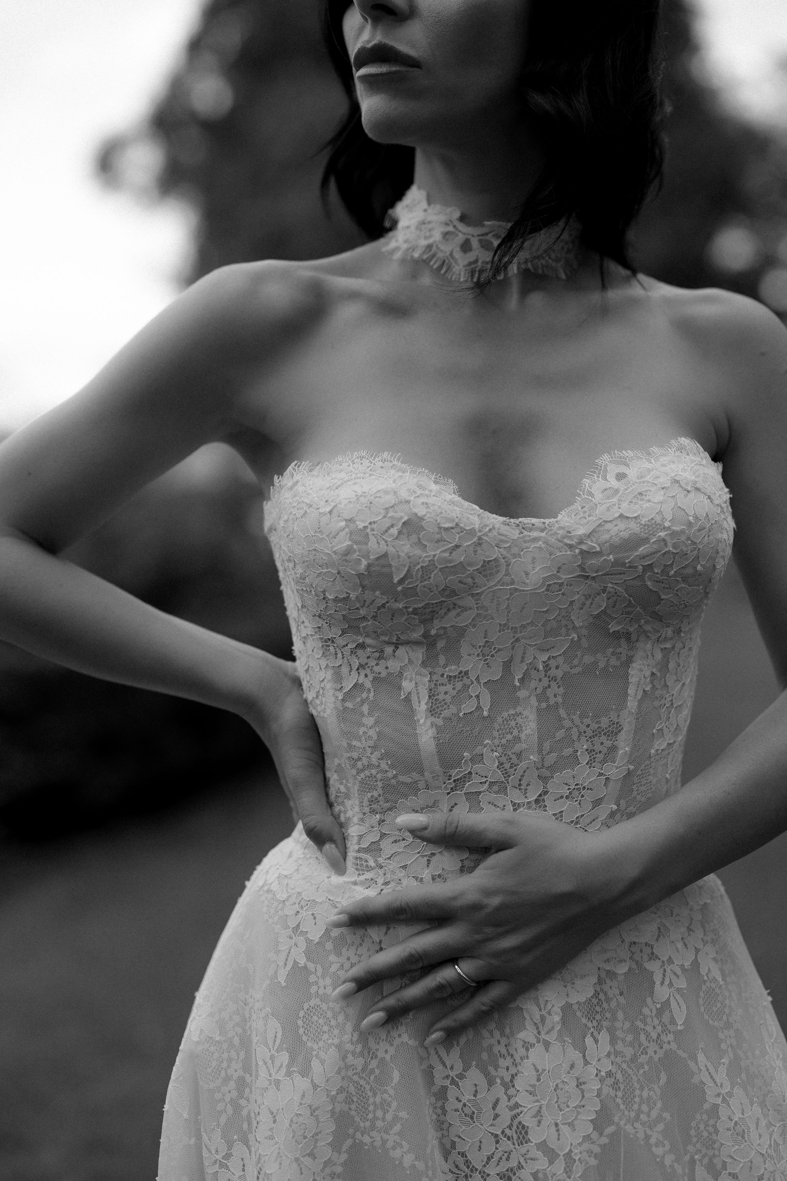 Black and white bridal photo of woman in strapless lace corset gown with choker necklace, hands on waist.