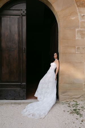Bride in strapless lace wedding gown with train stands in arched stone doorway of historic venue.