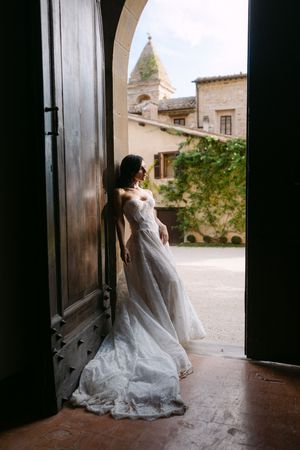 Elegant bride in flowing lace wedding gown standing in arched doorway of historic stone villa.