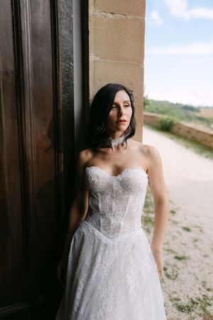 Elegant bride in strapless lace wedding gown leaning against stone wall outdoors.
