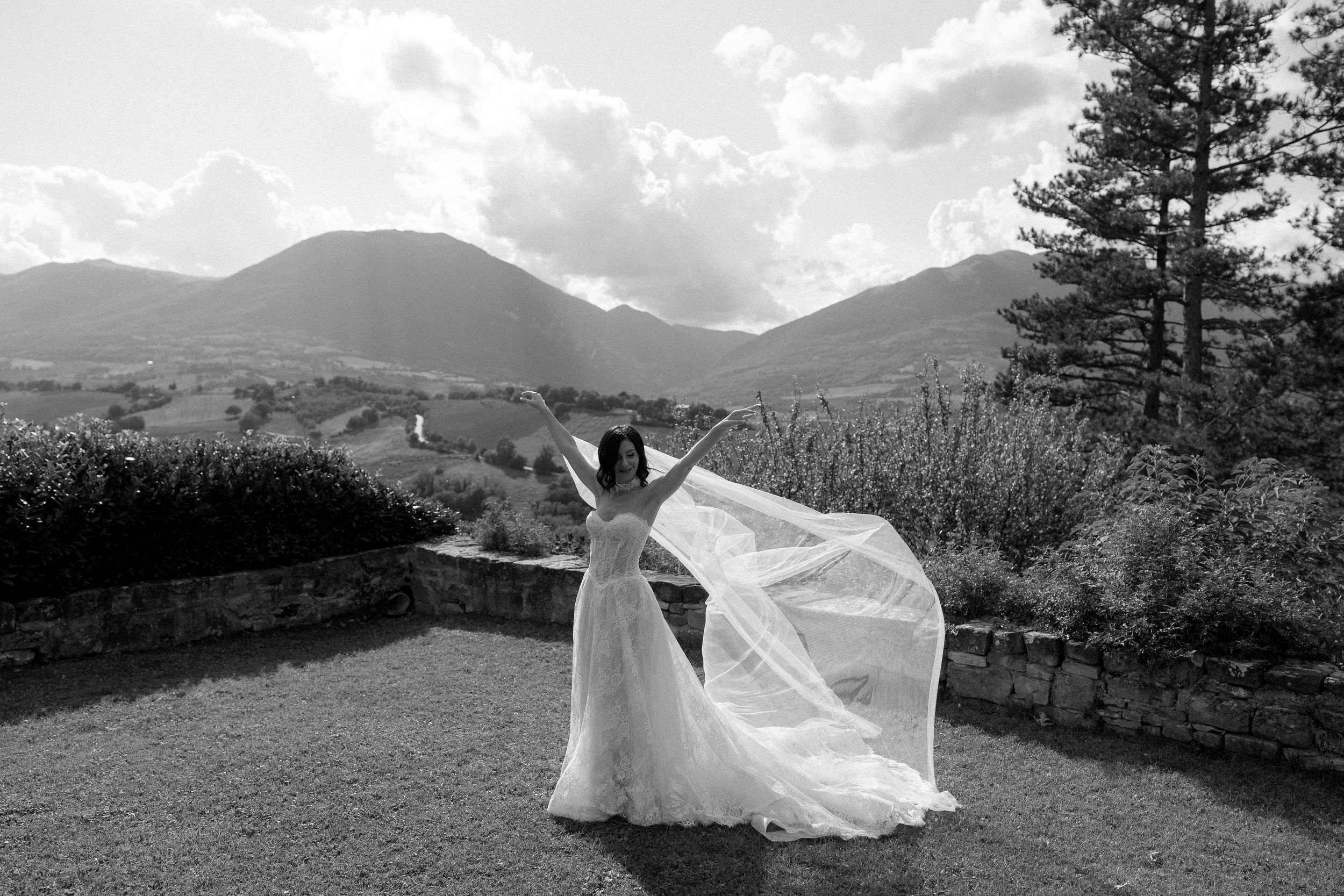 Black and white photo of a bride in a flowing gown with veil billowing, standing before a scenic mountain landscape.