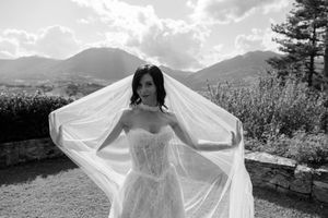 Bride in strapless lace gown holds flowing veil with mountains in background, black and white wedding photo.