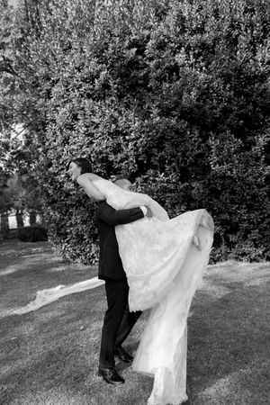 Groom lifts bride in flowing lace gown outdoors with lush green trees in black and white photo.