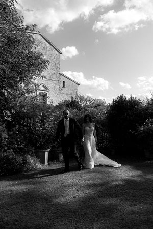 Black and white photo of a bride and groom walking in a garden beside a historic stone building on their wedding day.