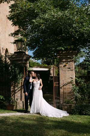 Bride and groom share a romantic kiss at an ornate stone garden gate surrounded by lush greenery on their wedding day.