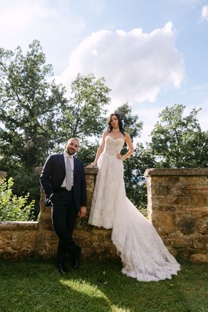 Bride in lace gown stands on stone wall beside groom in navy suit outdoors at elegant wedding venue.