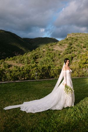 Elegant bride in a lace gown with long train poses in a green hillside vineyard under dramatic clouds.