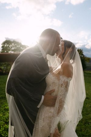 Bride and groom sharing a romantic kiss under a veil with sunlight glowing behind them outdoors.