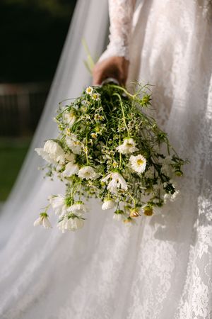 Bride holding cascading wildflower bouquet with white daisies against flowing lace wedding gown.