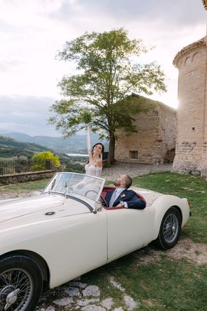 Bride and groom pose with vintage white convertible car beside historic stone tower at sunset.
