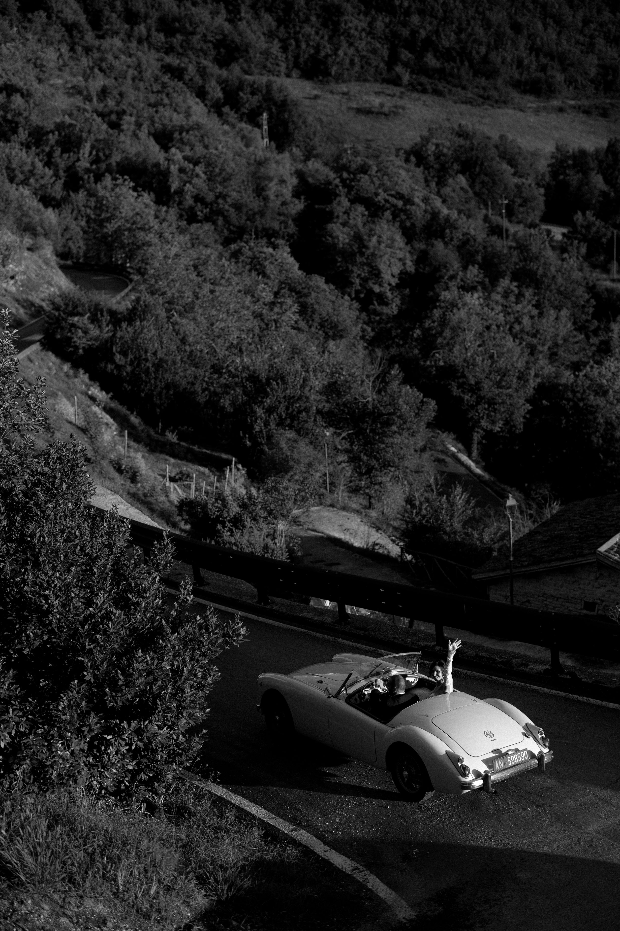 Newlyweds celebrate in a classic vintage convertible car driving through scenic countryside roads.