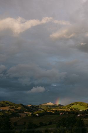 Dramatic stormy sky with rainbow over rolling green Italian countryside hills.