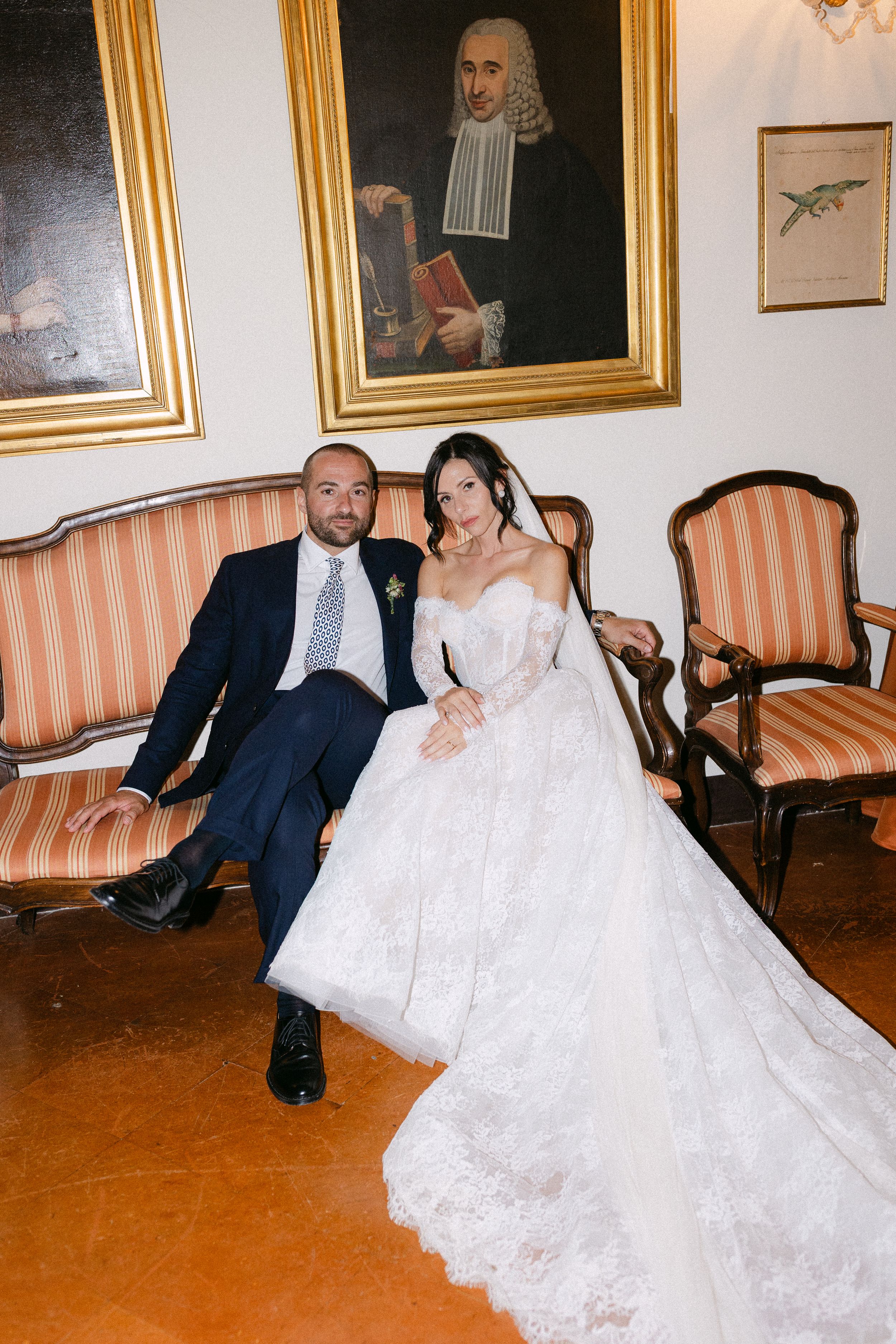 Bride in lace ball gown and groom in navy suit seated on striped sofa beneath classic portrait paintings.