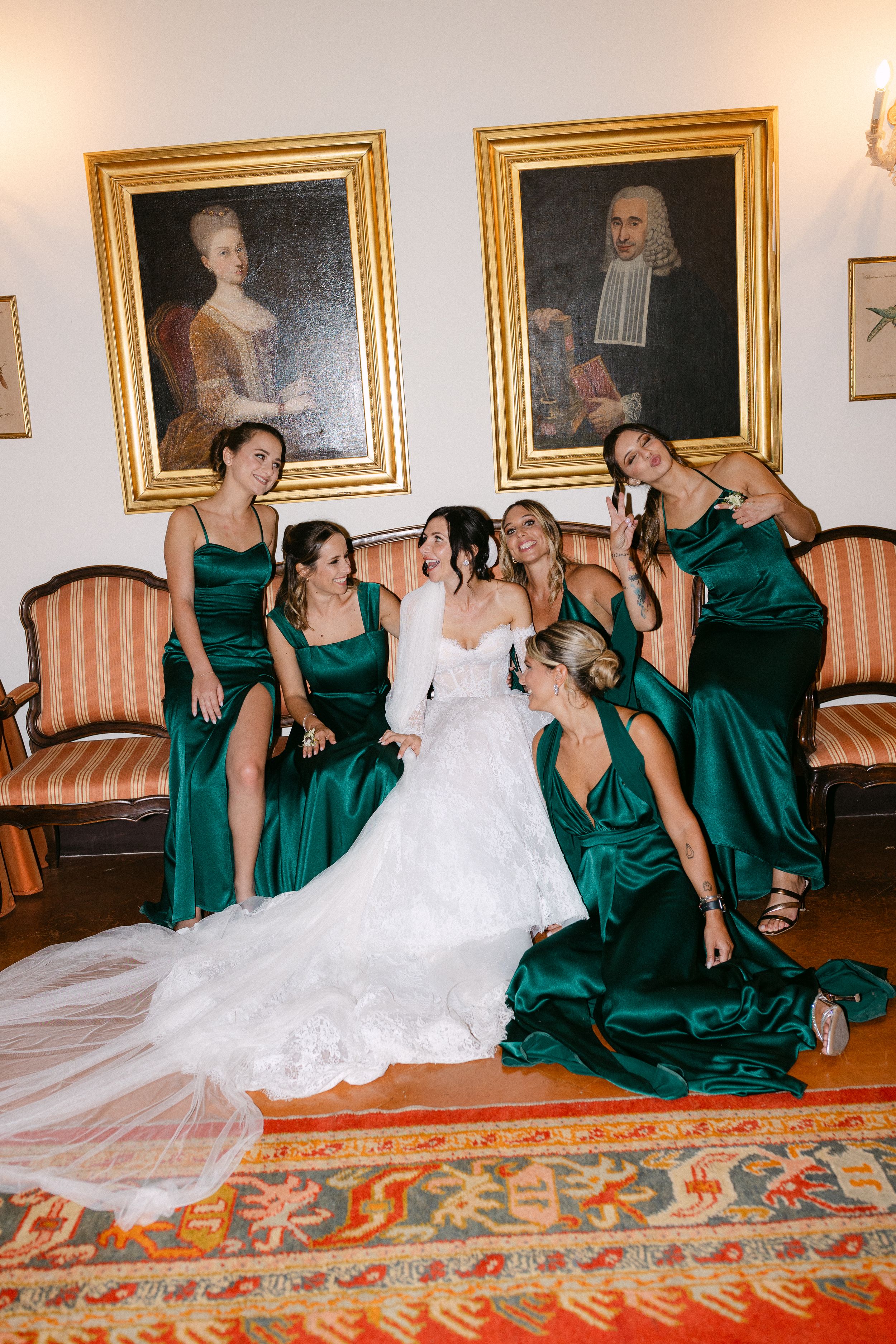 Bride in white gown poses with five bridesmaids in emerald green satin dresses before gold-framed portraits.