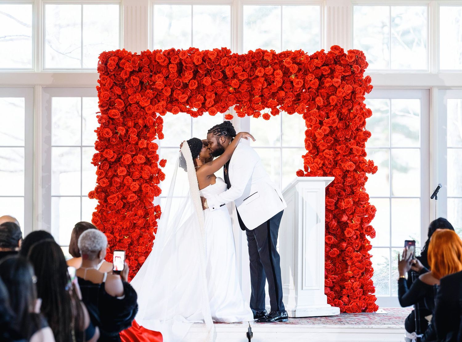 Bride and groom share first kiss under stunning red rose floral arch at elegant indoor wedding ceremony. Deerfield Golf Club Newark De