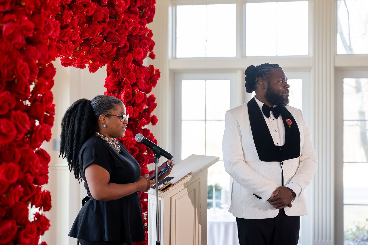 Woman speaking at podium beside man in white tuxedo jacket at elegant wedding ceremony with red rose floral arch. Deerfield Golf Club Newark