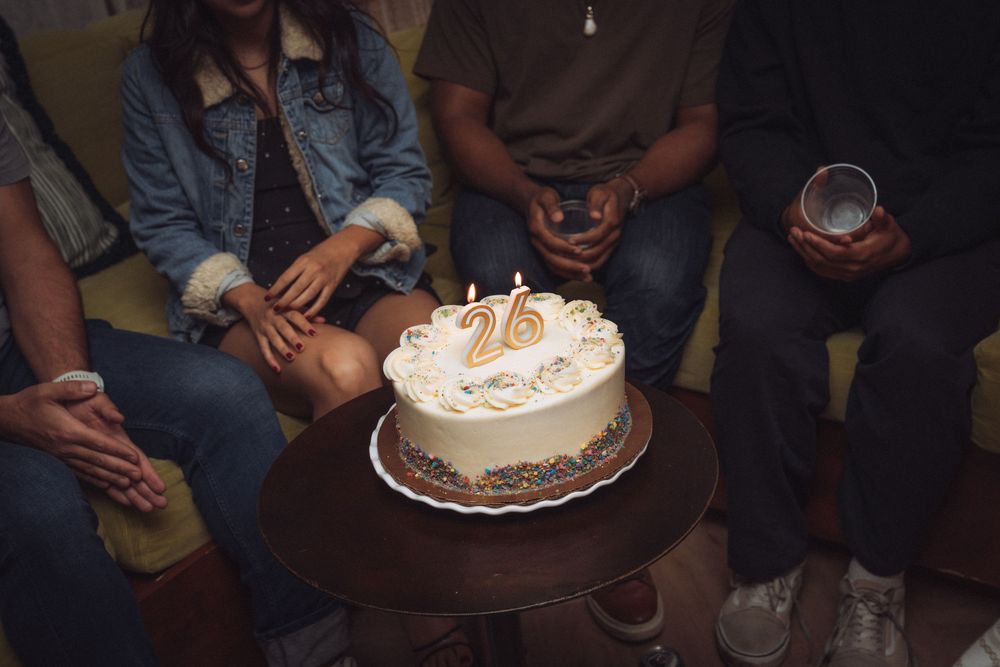 Friends sitting together celebrating 26th birthday with white frosted cake and candles.