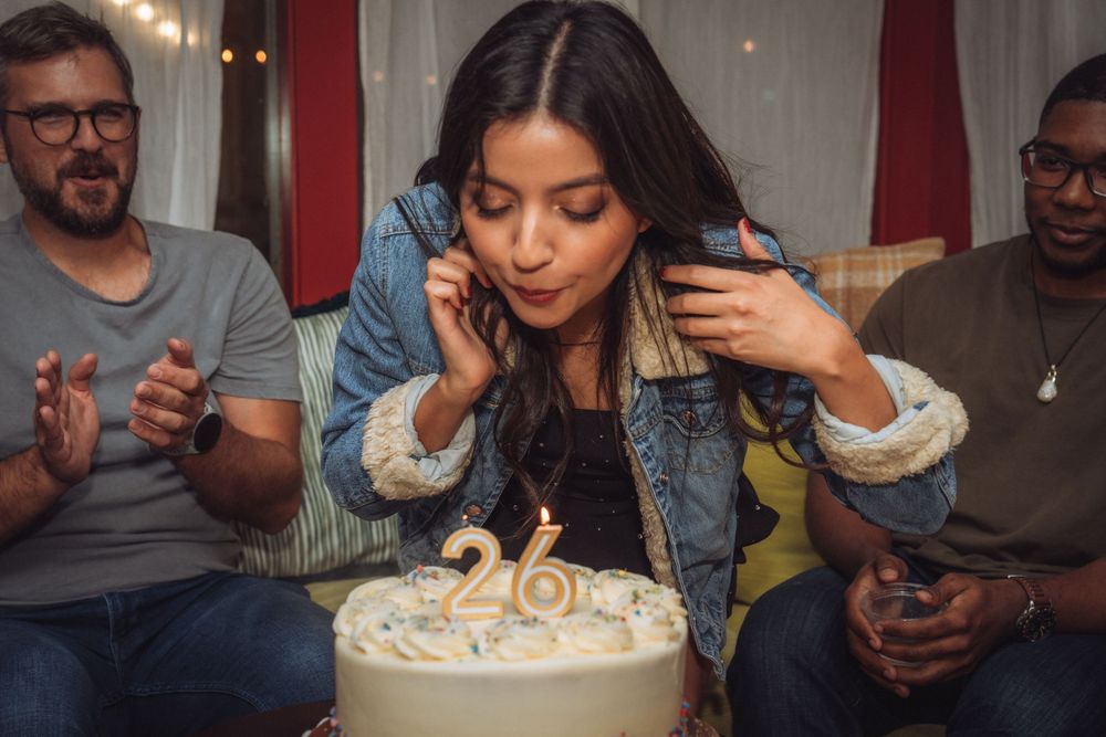 Woman blowing out candles on a 26th birthday cake surrounded by friends at a cozy indoor celebration.