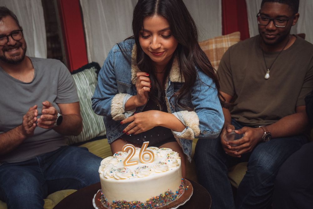 Woman blowing out candles on a 26th birthday cake surrounded by smiling friends at a party.